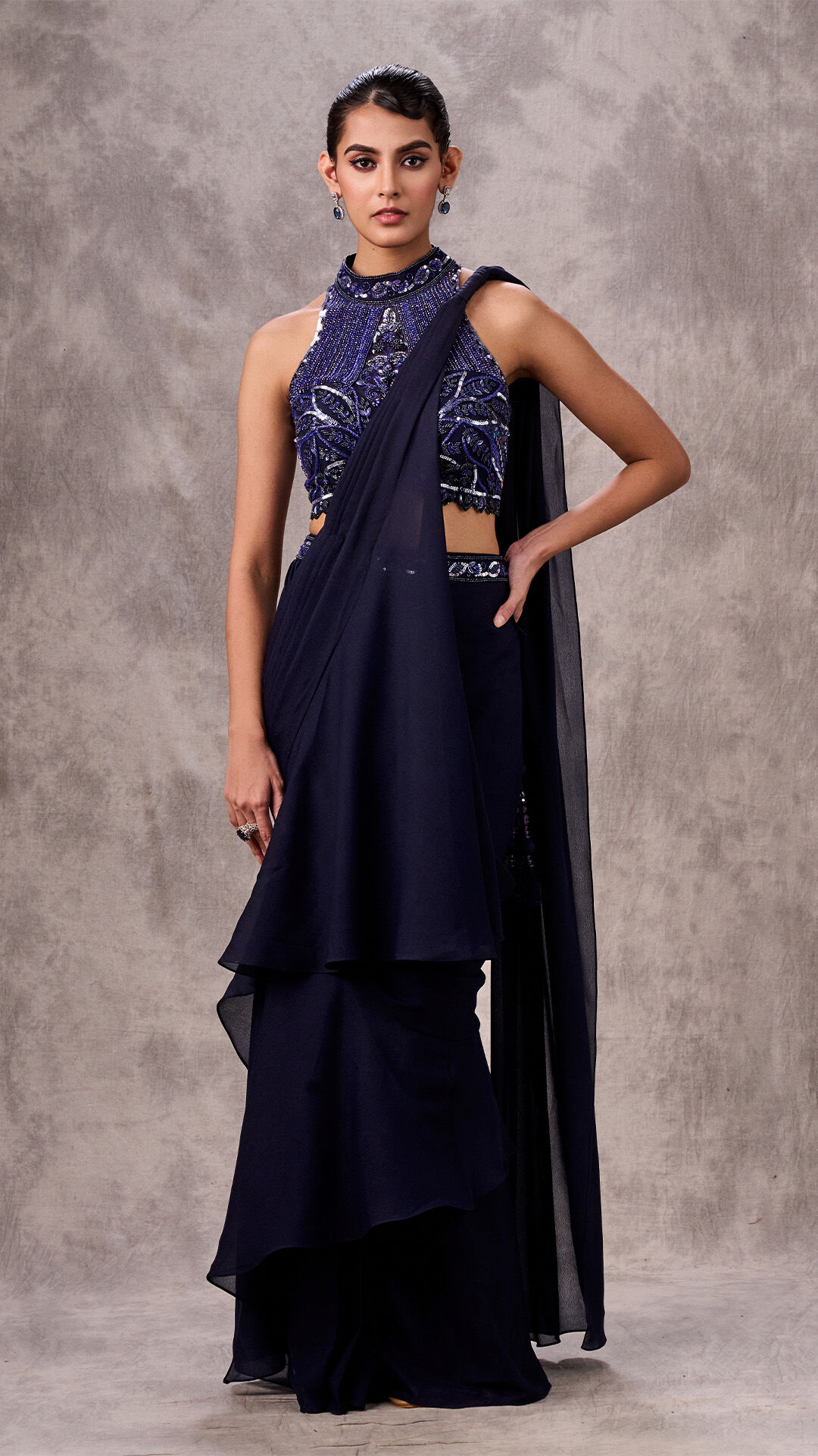 Woman modeling a pre-draped dark blue saree with a ruffled skirt and heavily embellished cut-out blouse, demonstrating a stylish blue ensemble.